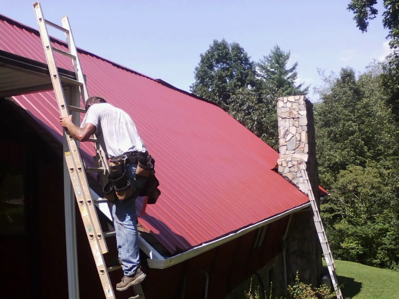 Roofer on a ladder beside a red metal roof and stone chimney — Soffit & Fascia Repair by Roofing Brothers in Chippewa Falls